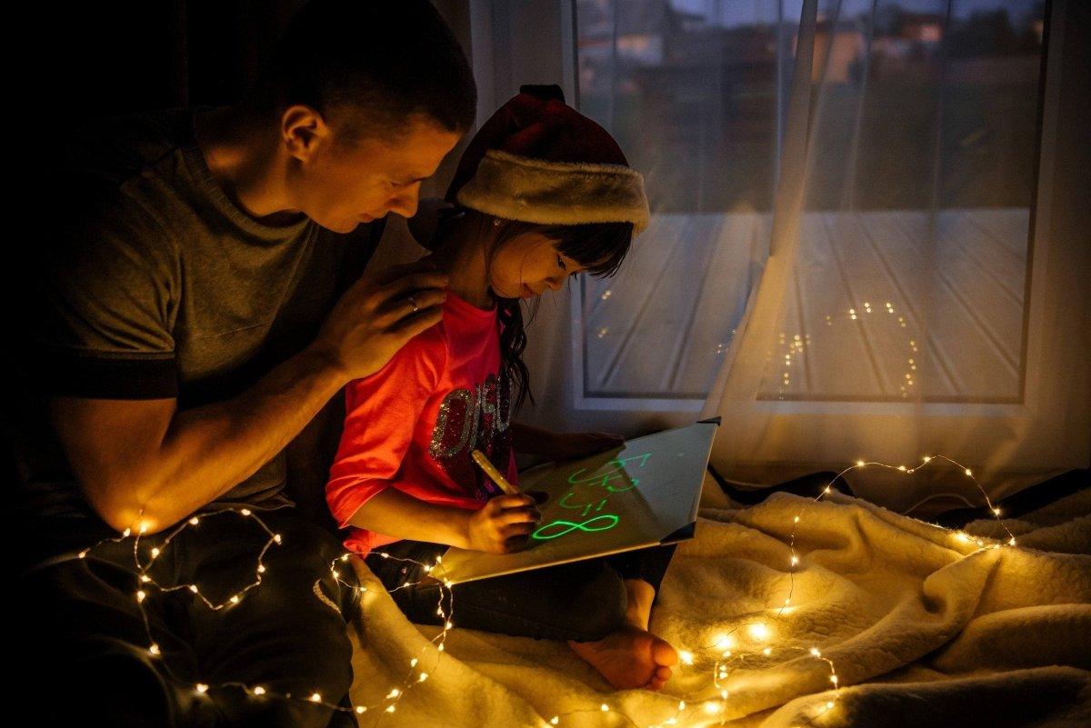 Adult helping child with glowing drawing pad in a cozy, illuminated setting with fairy lights.