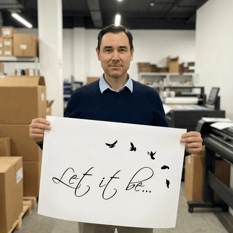 A man in a print workshop holds a white sheet with the words Let it be and black silhouettes of flying birds.