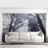A dramatic misty forest wallpaper mural covers the entire living room wall, depicting tall, shadowy trees disappearing into fog. The grayscale tones create a moody, immersive atmosphere behind a modern beige sectional sofa and minimalist coffee table.
