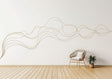 A large, abstract gold swirl line decal flows across a white wall in a minimal modern interior. A rattan chair with a white cushion sits below the decal, next to a small potted plant on a light wooden floor, creating a serene and contemporary atmosphere.