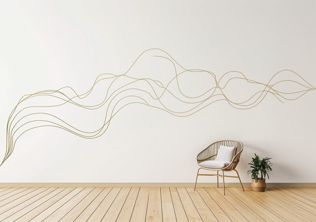 A large, abstract gold swirl line decal flows across a white wall in a minimal modern interior. A rattan chair with a white cushion sits below the decal, next to a small potted plant on a light wooden floor, creating a serene and contemporary atmosphere.