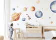 A child’s room with XXL watercolor wall decals of the solar system, including planets, stars, and a rocket. The child wears an astronaut helmet, standing near a light wood table and bed, in a softly lit, modern nursery space.