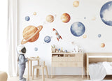 A child’s room with XXL watercolor wall decals of the solar system, including planets, stars, and a rocket. The child wears an astronaut helmet, standing near a light wood table and bed, in a softly lit, modern nursery space.