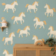A nursery wall featuring twelve pastel unicorn decals with golden manes and horns arranged over a teal background, above a wooden dresser with boho-style decor.