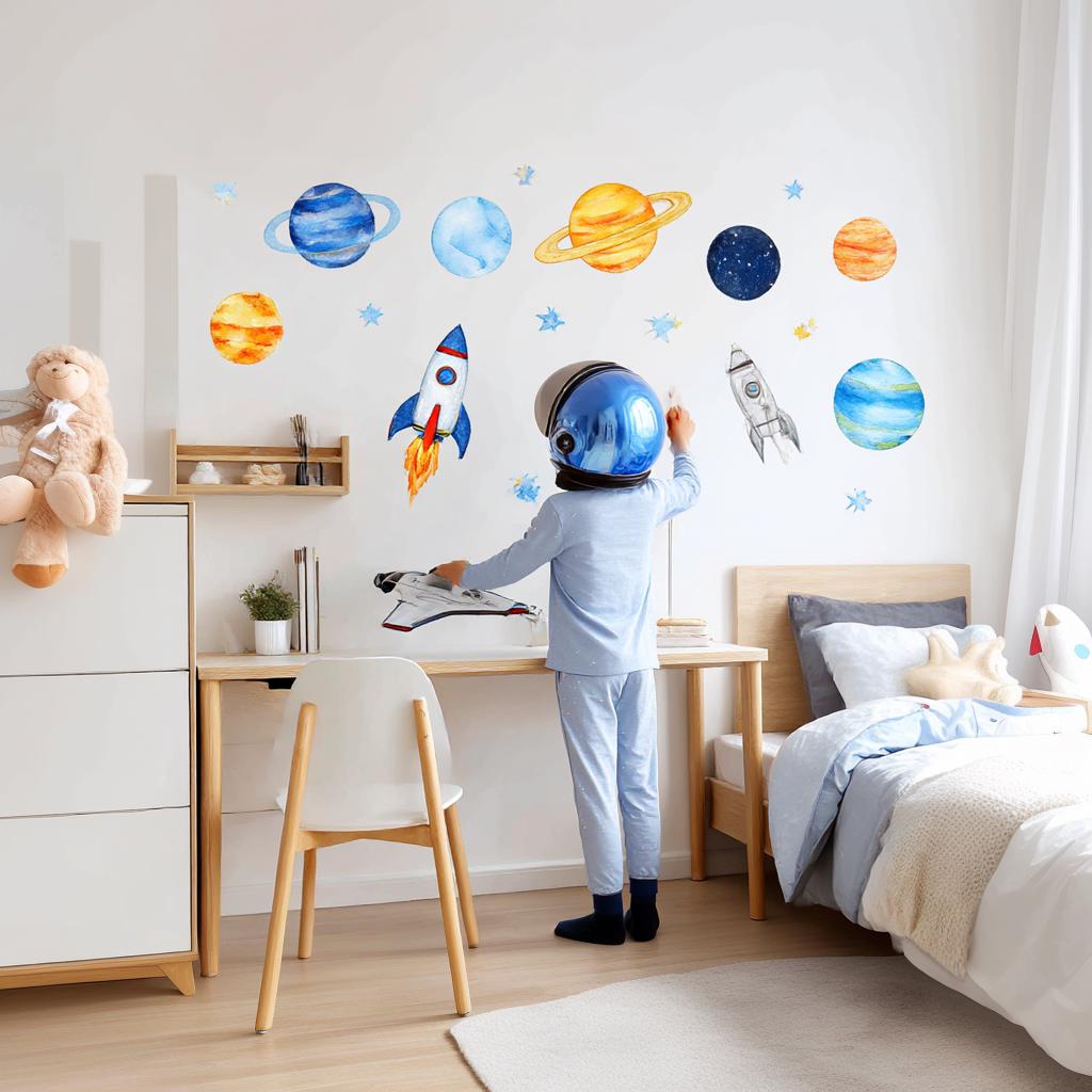 A child's bedroom with bright watercolor space wall decals, including planets, rockets, and stars in vivid blue, yellow, and orange hues. A child in an astronaut helmet plays near a desk and bed in a cozy, modern space-themed room.