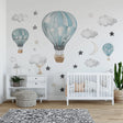 A dreamy nursery room with hot air balloon wall decals in soft blue tones, surrounded by watercolor clouds, stars, and a crescent moon. The wall art complements the white crib and wooden floor in a modern, cozy setting.