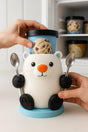 A 3D printed polar bear ice cream holder with a bright orange nose, holding two metal spoons in its paws, with a pint of ice cream on top and a built-in handle on the back. The bear sits on a blue base on a kitchen counter, with a freezer in the background.