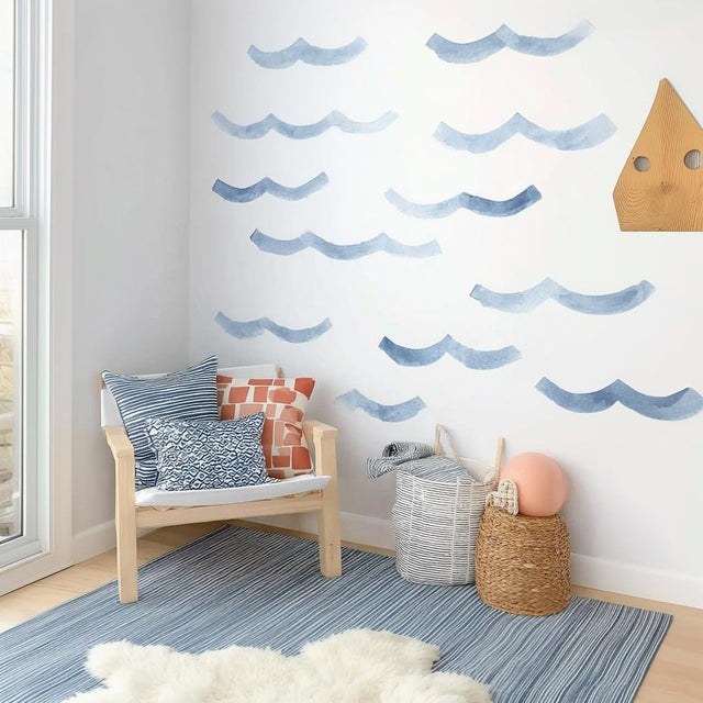 Watercolor wave wall decals in varying shades of blue arranged on a white wall above a cozy chair and storage baskets in a coastal-themed kids&#39; room.
