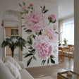 A bright, elegant living space with a large wall mural featuring soft pink watercolor peonies and green leaves. The floral design fills an accent wall between a cozy sitting area and a natural wood dining space, adding romantic charm and vibrancy.