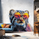 Printed vinyl wall decal of a Pallas's cat (manul) wearing large colorful sunglasses, with detailed fur in vivid colors. The decal is cut to shape and applied to a white indoor wall behind a modern sofa and interior elements.