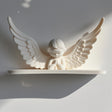 3D printed wall shelf with a minimalist praying angel figure and wide wings, matte white PLA, used as a small floating ledge on a light wall.