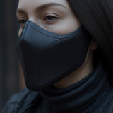 Close-up of a woman wearing a matte black 3D printed half-face mask with sleek curved lines, styled with a dark turtleneck against a soft background.