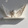 3D printed wall shelf with a minimalist praying angel figure and wide wings, matte white PLA, used as a small floating ledge on a light wall.
