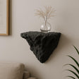 A black 3D printed wall-mounted shelf with a rugged, rock-like texture and triangular shape, displayed in a minimalist interior. The shelf holds a round glass vase with dried ornamental grass, and is mounted above a beige couch against a soft-toned wall.