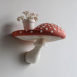 3D printed wall shelf shaped like a large red and white amanita mushroom with a flat spotted cap and sculpted gills, used as a small floating ledge holding a vase of flowers.