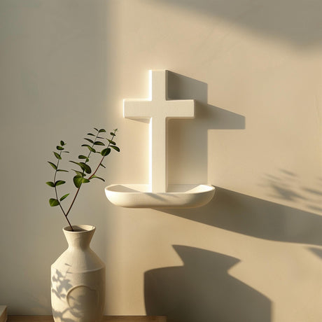 3D printed minimalist cross wall shelf in matte light PLA, with a small rounded ledge below the cross, mounted on a softly sunlit wall above a vase.