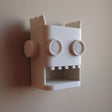 White 3D printed robot head wall organizer with an open mouth storage niche, made of matte PLA and mounted on a plain wall in soft natural light.