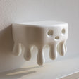 3D printed wall shelf shaped like a rounded dripping blob with a small face on the front, mounted on a light textured wall.