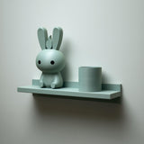 3D printed wall shelf with a slim back-edged ledge holding a rounded bunny character and a cylinder pot, mounted on a light-colored wall.