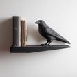 3D printed wall shelf with an integrated raven figure standing beside a row of books on a slim floating ledge against a light wall.