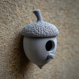 3D printed acorn-shaped birdhouse with round entrance and small cylindrical perch made of matte PLA plastic, mounted on a textured exterior wall.