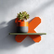 3D printed geometric X form wall shelf in orange with a green top, holding a potted plant, displayed as modern minimalist wall decor