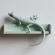 3D printed matte PLA wall shelf with an integrated gecko figure stretched along the ledge, its long tail hanging down against a light wall.