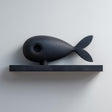 3D printed matte PLA wall shelf with a stylized fish figure resting on a slim rectangular ledge, mounted on a light-colored wall.