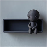 3D printed dark matte PLA wall shelf with an integrated stitched doll figure sitting on the edge against a light wall.