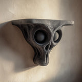 3D printed dark gray alien skull wall shelf with large hollow eye sockets and a flat floating ledge, mounted on a warm light textured wall.