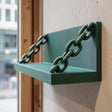 3D printed matte green wall shelf with integrated chain supports, mounted on a light textured wall near a window in a modern interior.