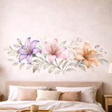 A horizontal watercolor illustration of three lilies in a row on a white background. From left to right: a light purple lily, a soft pink lily, and a warm peach lily. The flowers are connected by a central horizontal stem with delicate, pale green and grey leaves.