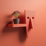 3D printed modern character wall shelf with abstract face design and integrated horizontal shelf, mounted on a warm terracotta colored interior wall.