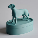3D printed oval pet ashes urn with a standing dog figurine on the lid, clean minimalist keepsake container with visible print texture on a studio background.