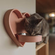 3D printed heart-shaped wall mounted pet bowl holder with a round bowl opening, shown with a cat eating from a metal bowl in a cozy home interior.