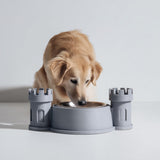 3D printed castle turret pet bowl holder stand with two tower sides and a central bowl opening, shown with a dog eating from a metal bowl in a studio setting.