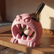 3D printed pink monster wine bottle holder with large teeth holding a wine bottle on a wooden shelf in warm sunlight.