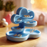 Light blue cloud-shaped jewelry organizer with layered trays and a round dish holding rings and earrings on a wooden table in warm indoor light.