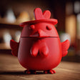 Red 3D printed chicken memorial urn with removable lid, rounded body, small wings, raised comb, beak, chest detail, and tiny feet on a wooden table.