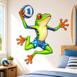 Bright green cartoon frog in blue floral shorts jumping while holding a blue-and-white volleyball, with orange toes and large red eyes on a white background.