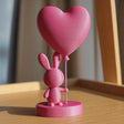 A close-up photograph of a 3D printed desktop figurine sitting on a wooden surface in warm sunlight. The cute figurine features a small minimalist rabbit standing on a circular base and holding a large heart-shaped balloon on a thin stick.