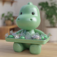 A close-up photograph of a 3D printed cartoon baby dinosaur figurine sitting on a wooden desk. The adorable creature holds a flat rectangular serving tray in front of it filled with wrapped candies, featuring a smooth rounded head, cute dark eyes, and a charming tabletop design.