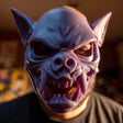 A close-up image of a person wearing a 3D printed, stylized fantasy monster mask. The mask features a feral creature design with pointed bat-like ears, an aggressive upturned snout, and a terrifying open mouth revealing large, sharp fangs.