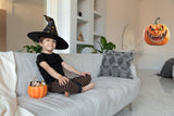 Child in witch hat sitting on couch with Halloween candy in pumpkin bucket and evil laughing pumpkin decal in background.