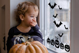 Child looking out window decorated with Halloween bats and pumpkins decals, celebrating the spooky season.
